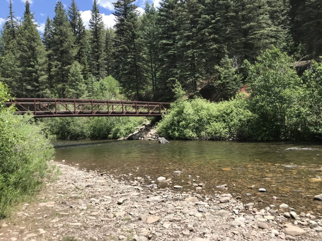 A rustic bridge crosses a river surrounded by lush trees.